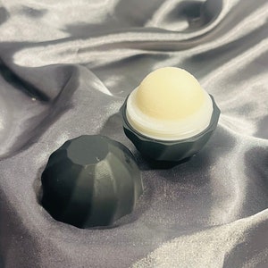 May include: A black and white lip balm in a black container with a geometric design. The lip balm is in a round shape and is in the open container.