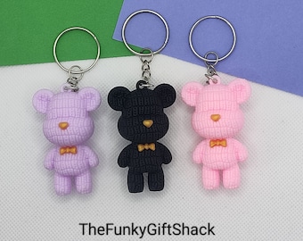 May include: Three teddy bear keychains, one pink, one black, and one light pink, with gold bow ties. The keychains are on a white background.