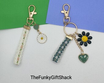 May include: Two keychains with gold-tone metal rings and charms. One keychain has a white daisy charm and a green heart charm. The other keychain has a black and yellow daisy charm, a green heart charm, and a gold chain. Both keychains have a white and green patterned ribbon.