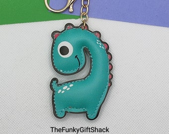 May include: A teal blue dinosaur keychain with a white eye and a pink mouth. The dinosaur is attached to a gold chain and a gold key ring.