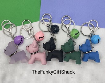 May include: Six colorful dog keychains with bells. Each keychain features a different color dog with a silver ring and a bell. The keychains are arranged in a row on a purple and green background. The text "TheFunkyGiftShack" is visible at the bottom of the image.