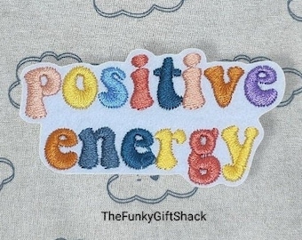May include: Embroidered iron-on patch with the words "positive energy" in rainbow colors on a white background.