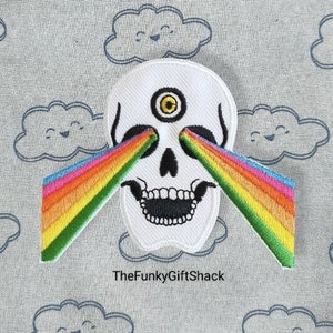 May include: White skull with a third eye in the centre of its forehead, shooting out rainbow beams of light. The skull is embroidered on a white fabric with a blue and white cloud pattern.
