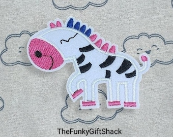 May include: White and black zebra applique with pink and blue details. The zebra is standing on all four legs with its head turned to the side. The applique is on a light gray background.