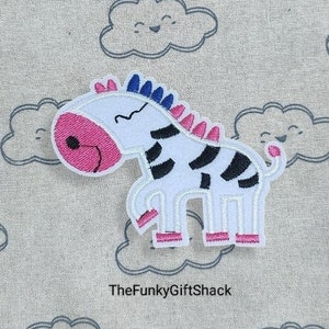 May include: White and black zebra applique with pink and blue details. The zebra is standing on all four legs with its head turned to the side. The applique is on a light grey background.