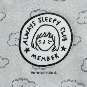 May include: White embroidered patch with black text that reads "Always Sleepy Club Member" and a cartoon drawing of a person with their eyes closed. The patch is on a grey fabric background with a cloud pattern.