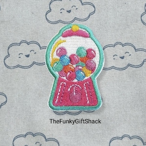 May include: An embroidered patch depicting a gumball machine with a pink base, a white top, and a green rim. The gumball machine is filled with colourful gumballs.