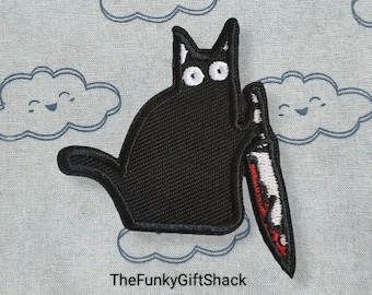 May include: Black embroidered patch of a cat holding a red and white knife. The cat has white eyes and is sitting on a light blue background with white clouds.