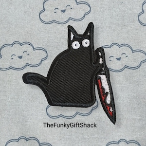May include: Black embroidered patch of a cat holding a red and white knife. The cat has white eyes and is sitting on a light blue background with white clouds.