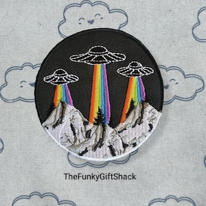May include: A black embroidered patch featuring three UFOs flying over a mountain range with rainbow beams of light. The patch is circular and has a white border.