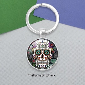 May include: A silver key ring with a round glass charm featuring a colourful sugar skull design. The skull is white with black outlines and has green, blue and purple floral details. The charm is surrounded by a silver ring.