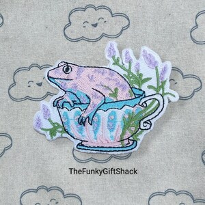 May include: Embroidered iron-on patch featuring a pink frog sitting in a blue and pink teacup with lavender flowers. The text "TheFunky GiftShack" is below the image.
