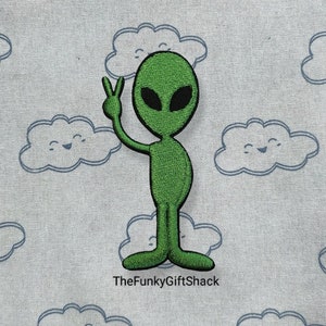 May include: Green alien with large eyes and a peace sign, embroidered on a white fabric with a cloud pattern.