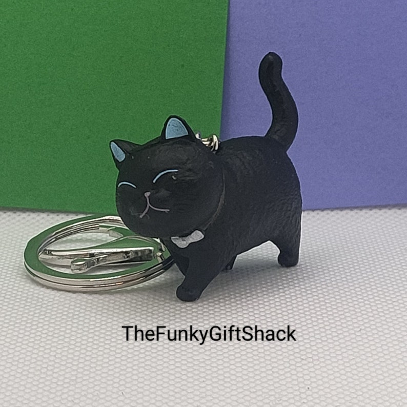 Puede incluir: Llavero de gato negro con una pajarita blanca. El gato est&aacute; sentado con la cola enroscada. El llavero est&aacute; unido a un anillo de metal plateado.