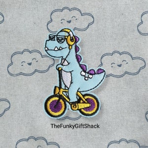 May include: A blue dinosaur wearing sunglasses and headphones rides a yellow bicycle with purple and yellow wheels. The dinosaur is embroidered on a light blue fabric with a white cloud pattern.