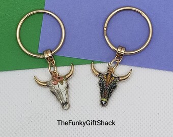 Puede incluir: Dos llaveros dorados con dijes de cráneo de toro. Un dije tiene un diseño floral y el otro tiene un diseño geométrico colorido. Los llaveros están sobre un fondo verde y morado. TheFunkyGiftShack está escrito debajo de los llaveros.