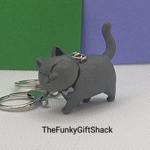 Puede incluir: Un llavero de gato gris con una pajarita, unido a un llavero plateado. El gato camina con la cola en el aire.