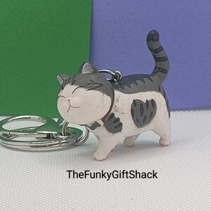 Puede incluir: Un llavero de gato blanco y gris con un parche en forma de coraz&oacute;n blanco y negro en el costado. El gato sonr&iacute;e y lleva un collar negro. El llavero est&aacute; unido a un llavero plateado.