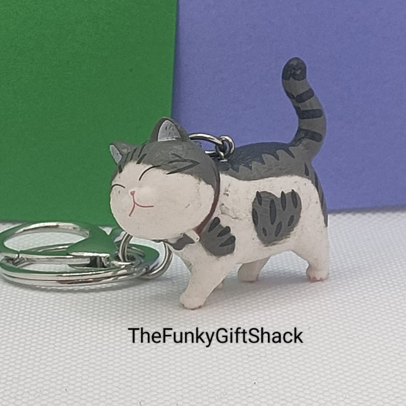 Puede incluir: Un llavero de gato blanco y gris con un parche en forma de coraz&oacute;n blanco y negro en el costado. El gato sonr&iacute;e y lleva un collar negro. El llavero est&aacute; unido a un llavero plateado.