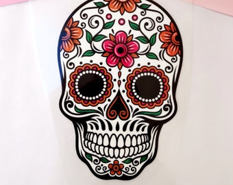 May include: A white sugar skull with black eyes, a red heart, and orange and pink flowers. The skull is decorated with green leaves and black swirls.