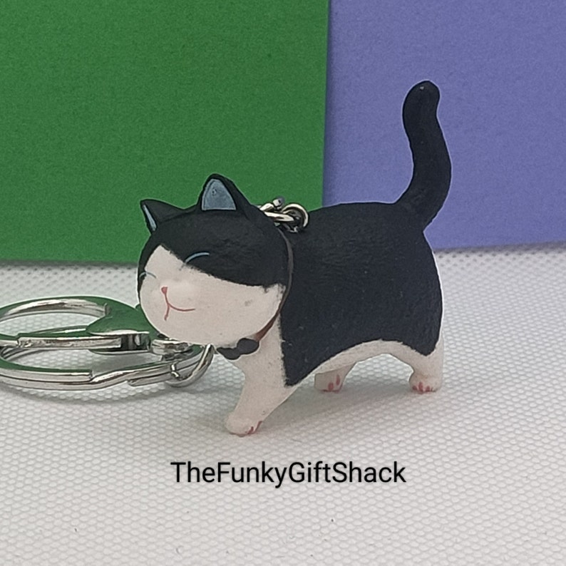Puede incluir: Llavero de gato blanco y negro con una cara sonriente. El gato est&aacute; de pie sobre sus cuatro patas con la cola enroscada. El llavero est&aacute; unido a un anillo de metal plateado.