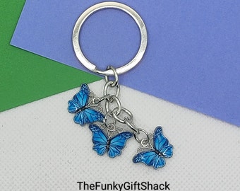 May include: A silver keychain with three blue butterfly charms. Each butterfly has a detailed wing design and a silver outline. The keychain is on a background of blue, green, and white.