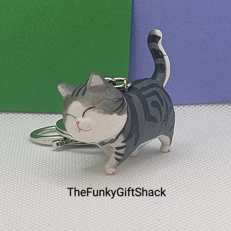 Puede incluir: Un llavero de gato gris y blanco con una cara sonriente. El gato est&aacute; de pie sobre sus cuatro patas con la cola enroscada. El llavero est&aacute; hecho de pl&aacute;stico y tiene un anillo de metal plateado.