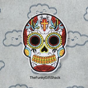 May include: White sugar skull embroidered patch with red, yellow and blue details. The skull has a cross on the forehead and intricate floral designs around the eyes.