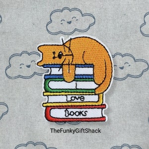 May include: Embroidered patch of an orange cat curled up on a stack of colourful books. The books have the words "Love Books" on them.