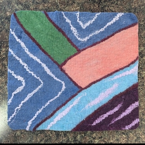 May include: A square, felted textile with a patchwork design. The design features a blue background with white zigzag lines, and sections of green, peach, light blue, and purple. The edges are trimmed with a dark red border. This item is likely a coaster or decorative mat.