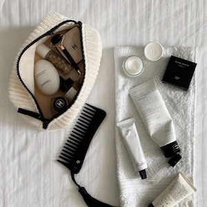 Pode incluir: Uma bolsa de tecido canelado cor de creme contendo um creme para as mãos Chanel em forma de ovo, um rolo facial e frascos. Ao lado, há um pente preto de dentes largos com borla, uma toalha branca e um tubo de creme Benton ceramide.