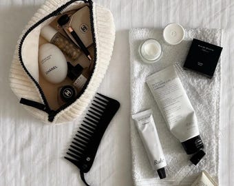 Puede incluir: Un neceser de tela acanalada color crema que contiene una crema de manos Chanel en forma de huevo, un rodillo facial y frascos. Al lado hay un peine negro de dientes anchos con borla, una toalla blanca y un tubo de crema Benton ceramide.