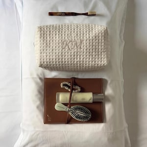Può includere: Una collezione di articoli per la cura personale che include uno spazzolino da denti tartarugato, un beauty case color crema con trama a nido d'ape e le lettere KM ricamate, un taccuino in pelle marrone, un elastico per capelli, una lozione e una spazzola.
