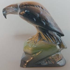 Op de afbeelding: Een glanzend keramieken beeldje van een adelaar in zittende houding. De vogel heeft een donkerbruin en zwart verenkleed met een witte kop en nek. Het beeldje staat op een groen en bruine rotsachtige basis met een gladde, reflecterende afwerking.
