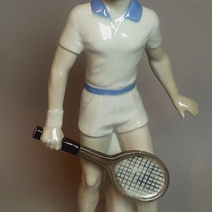 Op de afbeelding: Een porseleinen beeldje van een tennisser met bruin haar, gekleed in een wit poloshirt met blauwe kraag, witte korte broek met blauwe riem en witte sneakers met gele sokken. Het figuur houdt een tennisracket met een zwart handvat vast.