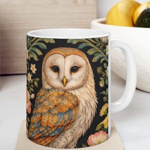 May include: White ceramic mug featuring a detailed illustration of a barn owl with a cream-colored face, orange and blue wings, and a black background with floral accents. The mug is on a beige coaster.