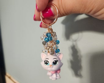 May include: A white cat charm with blue eyes and a pink bow, attached to a gold key ring with blue and clear crystal beads.