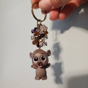 May include: A gray cartoon animal keychain with large eyes and a brown nose. The keychain has a cluster of purple, clear, and brown beads attached to a gold key ring.