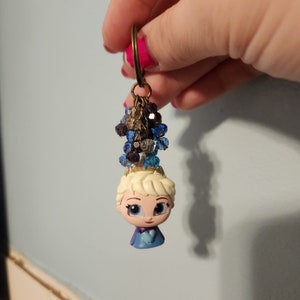 Puede incluir: Una figurita azul y blanca de una princesa con una corona y un vestido azul. La figurita est&aacute; unida a un llavero con un racimo de cristales azules y transparentes.