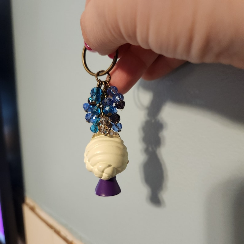 Puede incluir: Un amuleto blanco y morado con cuentas azules y transparentes que cuelgan de un llavero dorado.