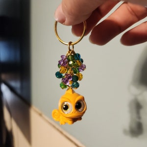 May include: A gold-colored keychain featuring a small, yellow chameleon charm with large eyes. The charm is accented with a cluster of colorful beads in shades of purple, green, blue, and yellow. The keychain is held by a gold ring.