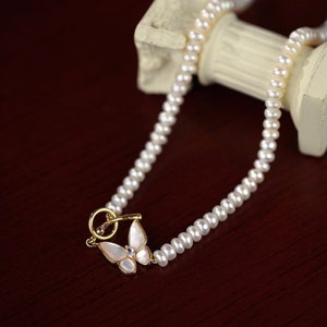 Puede incluir: Un delicado collar de perlas con un dije de mariposa dorado. El collar está hecho de pequeñas perlas blancas y tiene un diseño simple y elegante.