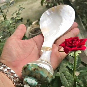 May include: A white mother of pearl spoon with a green and brown shell handle. The spoon is held in a hand with a silver chain bracelet.