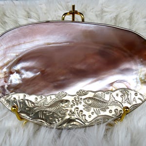 Puede incluir: Un plato decorativo ovalado con una superficie nacarada en tonos marrones y rosas. El plato tiene un borde plateado ornamentado con diseños de peces y flores. Está sostenido por detalles metálicos dorados.