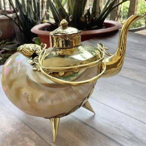 May include: A gold teapot with a shell-shaped body and a decorative lid. The teapot has a long spout and a handle with a decorative design. The teapot is sitting on a wooden surface.