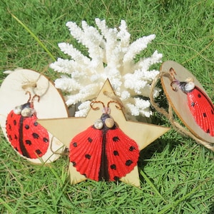 May include: Three wooden ornaments shaped as a star and two circles, each featuring a ladybug design made from seashells. The ladybugs are painted red with black spots and have white shell accents.