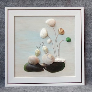 Op de afbeelding: Een ingelijst kunstwerk van schelpen met een witte achtergrond. Het kunstwerk bevat drie kleine schelpen met witte en bruine accenten, gerangschikt om op slakken te lijken. De slakken bevinden zich op een bed van twee grotere schelpen, die bruin en groen zijn. Er zijn vijf witte schelpen gerangschikt om op bloemen te lijken met groene en bruine accenten.