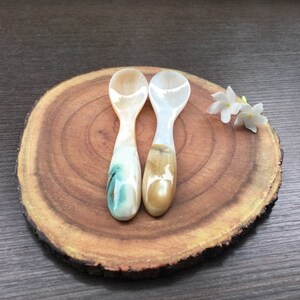 May include: Two small spoons made of mother of pearl with a light brown and green swirl pattern. The spoons are on a wooden surface.