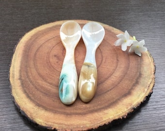 May include: Two small spoons made of mother of pearl with a light brown and green swirl pattern. The spoons are on a wooden surface.