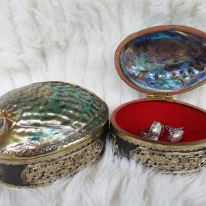 May include: An open, oval-shaped jewelry box with a red interior and a pair of earrings. The box is made of a shell with iridescent green and blue hues, and is trimmed with gold-colored metal. The box is next to a closed matching box.